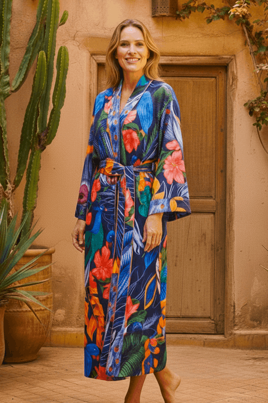 Woman looking to camera wearing the Lunar Bloom exclusive print silk-cotton robe in navy with peacock and parrot print, long length, styled in a Moroccan courtyard