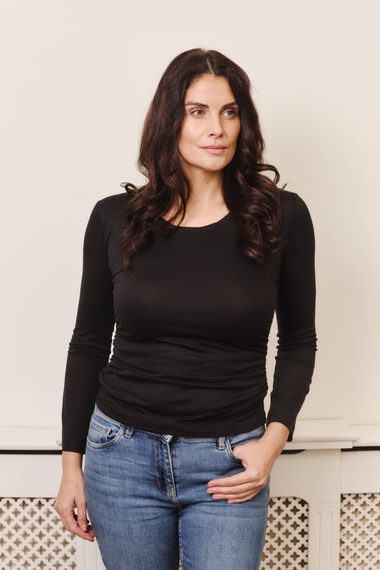 Woman wearing the Alya top, which has a scoop neck, long sleeves and ruching on the body below the bust area. Fabric is 96% TENCEL™ Lyocell, 4% Elastane - a stretch jersey, with a silky soft touch and fluid drape.